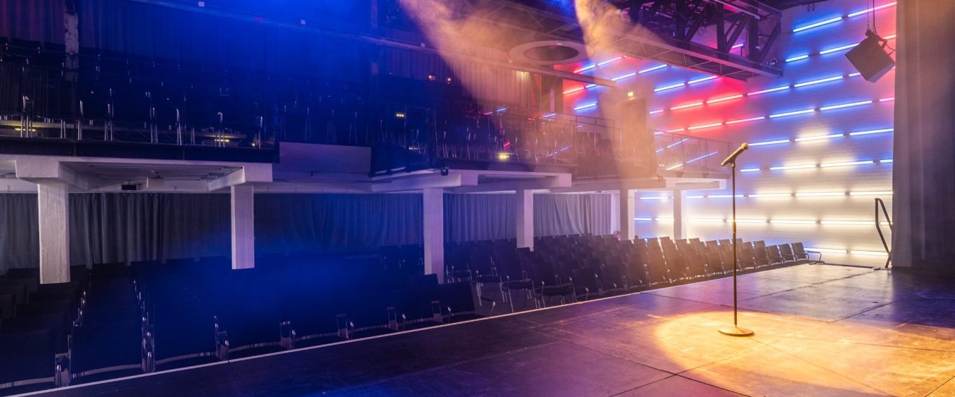 Großer Saal im Kulturwerk Blick von der Bühne. Leere Reihen von schwarzen Stühlen in einem modernen, bunten Veranstaltungsraum.