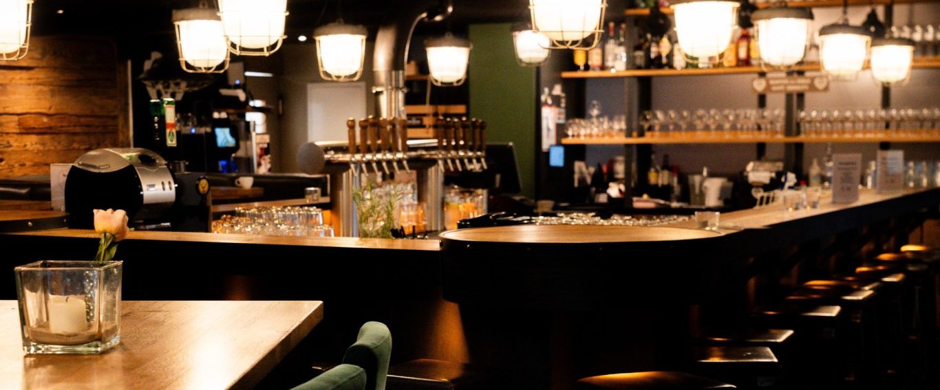 Tresen in der Hopfenliebe Moderner Tresen mit Holzdetails, Barhockern, beleuchtet von warmen Lampen und einem großen Gläserregal im Hintergrund.